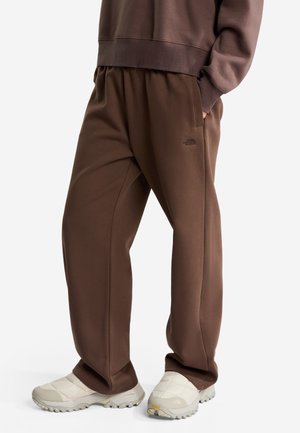 Person, die lockere braune Sweatpants und einen passenden Sweatshirt mit weißen Slip-On Outdoor-Schuhen trägt, steht mit einer Hand in der Tasche.
