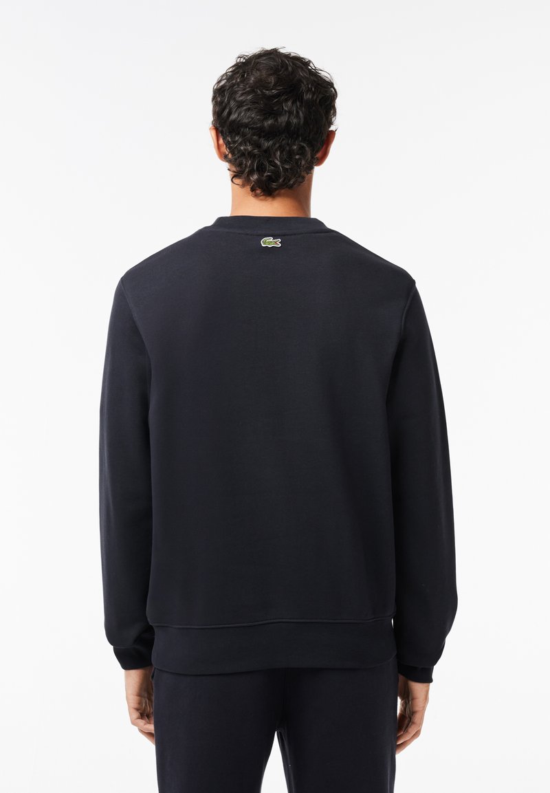 Hombre con cabello rizado oscuro vistiendo un sudadera negra y pantalones, mostrado de espaldas contra un fondo blanco liso.