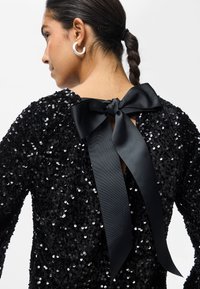 Mulher com cabelo trançado, usando grandes brincos de argola brancos e uma blusa preta de lantejoulas com um grande laço de cetim amarrado nas costas.
