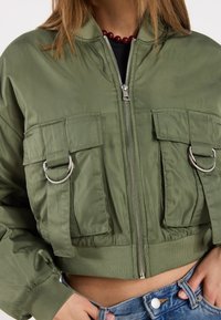 Skraćena bomber jakna, maslinasto zelena, od najlonskog materijala, sa dva prednja džepa sa srebrnim detaljima i zatvaranjem na patent. Teksturisani rukavi na donjem rubu.