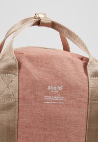 Sac à dos en tissu rose avec des accents dorés métalliques, doté d'une poignée supérieure et d'un logo en texte. Fermeture éclair et poignées texturées pour plus de durabilité.