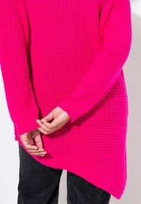 Helles pinkes, strukturiertes Strickpullover mit lockerer Passform, gerippten Bündchen und asymmetrischem Saum. Über schwarzen Hosen getragen, die Hände korrigieren den Saum.