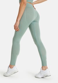 High-waisted leggings in light green, featuring a smooth texture, contrasting blue stitching, and a snug fit. Worn with white sneakers.