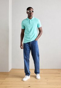 Polo shirt bleu clair à manches courtes avec un tissu texturé, associé à un jean en denim bleu ample et des baskets blanches. Lunettes de soleil carrées blanches.