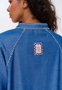 Džínová košeľa s kontrastným bielym prešívaním, so štvorcovou štítkom s červenobielym károvaným okrajom a textom "Ver v svoj svit."