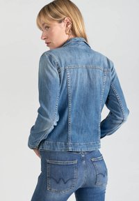 Giacca di jeans blu con un design aderente, cuciture a contrasto e polsini con bottone. Indossata con jeans di jeans abbinati caratterizzati da accenti sulle tasche.