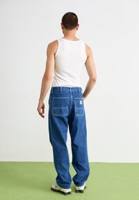 Blue denim pants with a relaxed fit, featuring two back pockets and a small logo patch. Worn with a white tank top and sneakers.