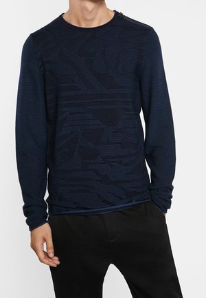 Uomo che indossa un maglione a maniche lunghe blu navy con motivi astratti e pantaloni neri, mani rilassate ai lati.