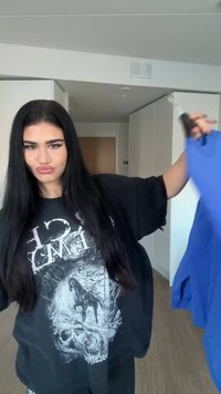 Mladá žena s dlouhými černými vlasy, oblečená v černém tričku s potiskem, drží modrý oděv v moderní bílé kuchyni nebo obývacím prostoru.
