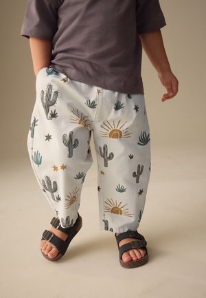 Kind draagt witte broek met cactus- en zonpatronen en zwarte sandalen, staat met één hand in de zak.