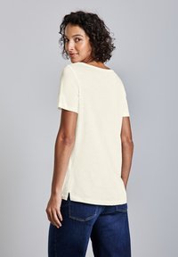 Kortærmet, off-white skjorte med scoop halsudskæring, sideskår og en afslappet pasform, parret med mørkeblå jeans.