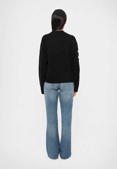 Črn pleten pulover z okroglim izrezom in rebrastim robom, kombiniran s svetlo modrimi razširjenimi kavbojkami. Gladek teksturni videz z minimalnimi detajli.