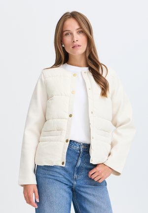 Femme portant une veste matelassée blanche avec des boutons-pression, un top blanc et un jean bleu, posant devant un fond clair uni.