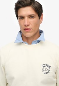 Sudadera crema con cuello redondo, textura suave y logotipo en azul marino. Camisa de color azul claro con cuello visible debajo, con detalles de botones.