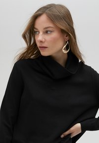 Svart tröja med kapuschong i stickat tyg och långa ärmar; modellen har accessoarer med stora, gyllene droppformade örhängen. Bakgrunden är neutral.