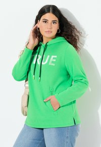 Felpa verde con "TRUE" stampato in bianco, cappuccio con coulisse e accenti neri e verdi, tasche laterali e spacco laterale, abbinata a jeans blu.