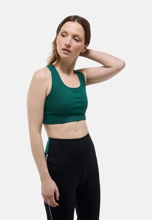 Femme à la peau claire et aux cheveux bruns, portant un brassard de sport vert et un legging noir, posant avec une main derrière la tête sur fond blanc.