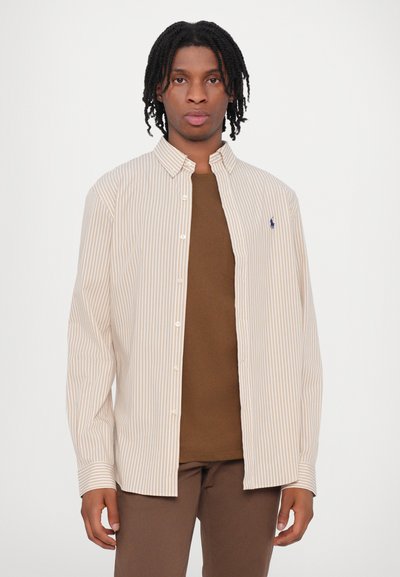 Dryžuota šviesiai ruda ir balta marškinėlių palaidinė su laisvu kirpimu, turinti apykaklę, ilgomis rankovėmis ir mažą logotipą krūtinėje.