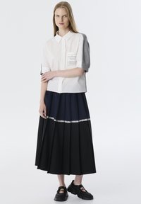 Camisa blanca de manga corta con detalle gris, bolsillo funcional, combinada con una falda plisada negra y azul marino con encaje. Se llevan zapatillas negras.