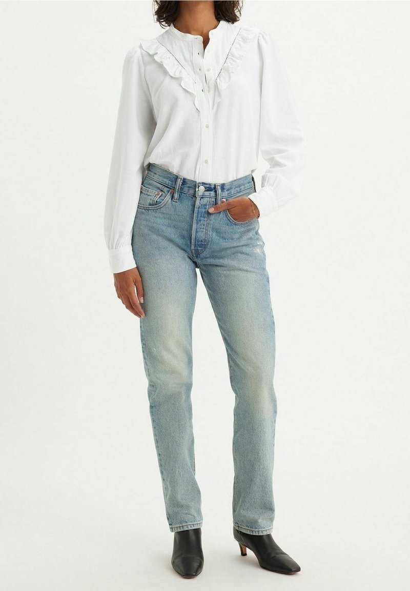Femme portant un chemisier blanc à col à volants, un jean droit bleu clair et des bottines noires à talons, posant avec une main dans la poche.