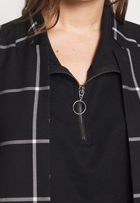 Gros plan sur une personne portant un haut zippé noir avec un tirette en anneau argenté et une veste noire à motif à carreaux blancs.