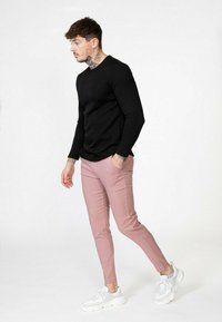 Chemise noire à manches longues côtelée associée à un pantalon rose clair ajusté et des baskets blanches, présentant un design minimaliste et des détails de tatouage subtils.