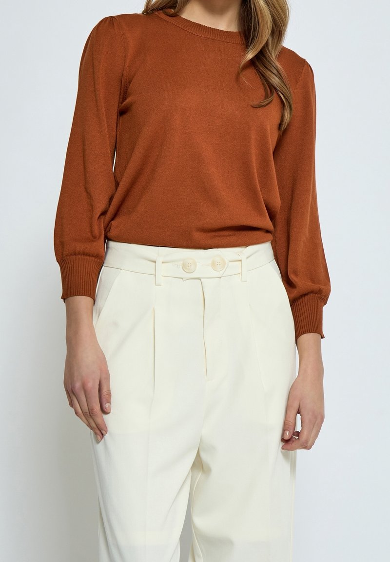 Femme portant un pull à manches longues couleur rouille et un pantalon crème taille haute avec deux boutons sur le devant, visible du cou jusqu’au milieu de la cuisse.