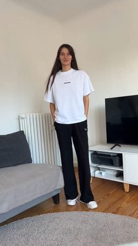 Balta per didelė marškinėliai su juodu tekstu, derinami su juodomis kelnėmis. Medinės grindys, pilkas sofa ir televizorius fone.