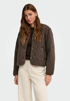 Jonge vrouw met lang bruin haar die een bruine fleecejas met knopen draagt over een donker topje en crèmekleurige hoge taillebroeken.