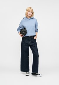 Felpa oversize blu chiaro, jeans a gamba larga blu scuro, marsupio nero, scarpe da ginnastica bianche e nere, con uno sfondo semplice.