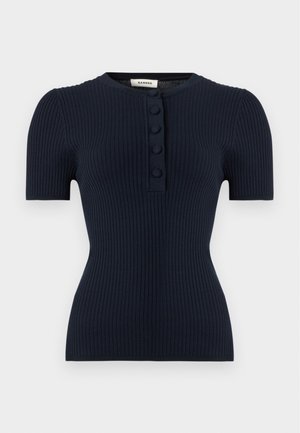 Femme portant un haut noir côtelé à manches courtes, un pantalon large bleu marine, des chaussures noires pointues et de grandes lunettes de soleil noires, debout sur fond blanc.