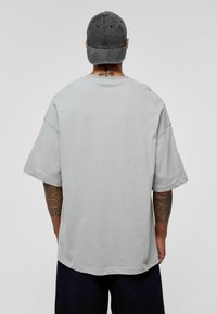 Grijze oversized T-shirt met korte mouwen, gemaakt van zachte katoen. Gedetailleerd achteraanzicht dat een tatoeage toont die contrasteert tegen de stof.