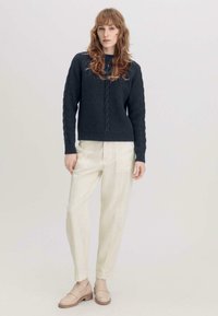 Dunkelgrauer gestrickter Pullover mit Zopfmuster, kombiniert mit hellbeigen Hosen und beigen Slippern, vor einem neutralen Hintergrund.