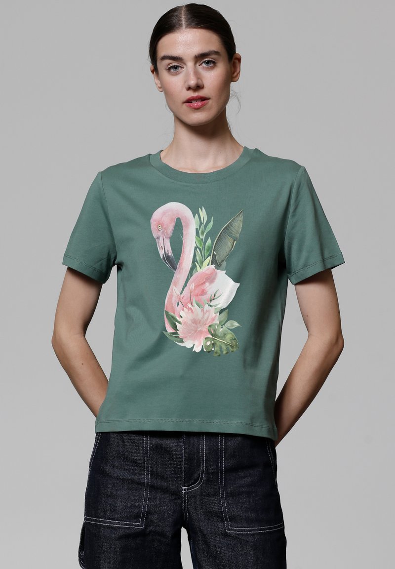 T-shirt en coton vert avec un design de flamant rose et de fleurs sur le devant. Manches courtes et col rond. Pantalons en denim foncé portés en bas.