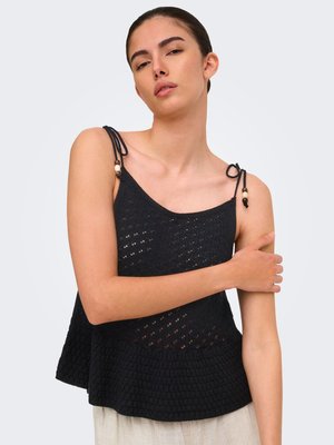 Jeune femme portant un haut en maille noir sans manches avec des bretelles à nouer et des perles, posant avec un bras croisé sur son corps.