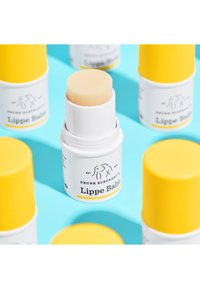 Drunk Elephant Lippe-Balm mit einem runden, hellgelben Stift in einem weißen Behälter, der einen gelben Deckel und Branding-Details aufweist. Heller blauer Hintergrund.