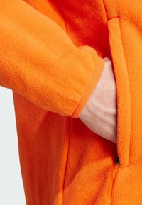 Orange fleecejakke med en blød tekstur, hvor en hånd er stukket ind i en sidelomme med en kontrastfarvet lynlås i orange.
