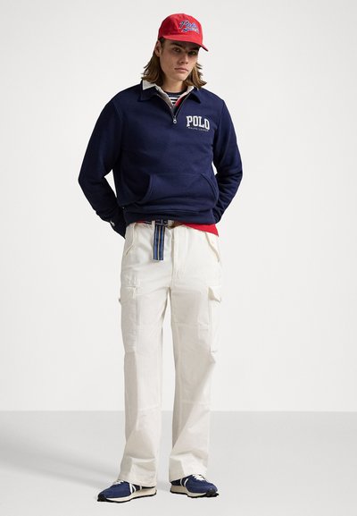 Marine fleece pullover med halvlange lynlås, hvide cargo bukser, rød kasket med logo og blå sneakers. Har skjorte med striber under.