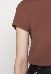 Haut marron à manches courtes avec une texture lisse et un subtil éclat, présentant une coupe décontractée et une couture simple le long des bords des manches.