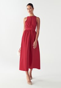 Sieviete, tērpusies sarkān bezmaku midi kleitā ar savilkta vidukļa daļu, tur baltu rokassomu un valkā beiges norādītās kurpes uz fona bez raksta.