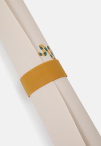 Deux tapis roulés de couleur crème maintenus par un élastique rond jaune avec le texte en relief "Lifewood". Des détails rayés verts et jaunes sont visibles.