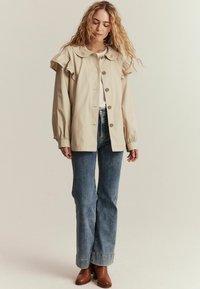 Beige jakke med bølgende ærmer og dekorativ krave, knaplukning, parret med højtaljede blå jeans og brune hælede støvler.