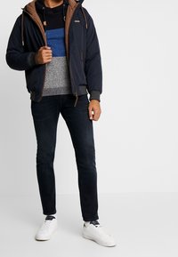 Veste matelassée bleu marine avec doublure en laine polaire marron, pull rayé gris et bleu, jean foncé et baskets blanches. Tissu lisse, coupe décontractée.