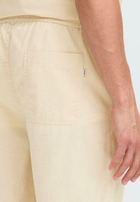 Lysbeige bomullsshorts med avslappet passform, som har en sidelomme og en liten merkelapp på baksiden. Myk tekstur og enkel design.