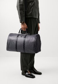 Sac de voyage à carreaux noirs avec des accents en cuir lisse et deux poignées. Il dispose d'une fermeture zippée et d'un logo en relief à l'avant.