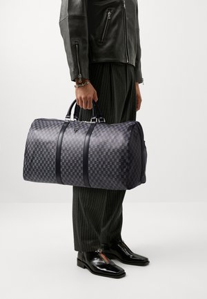 Sac de voyage à carreaux noirs avec des accents en cuir lisse et deux poignées. Il dispose d'une fermeture zippée et d'un logo en relief à l'avant.