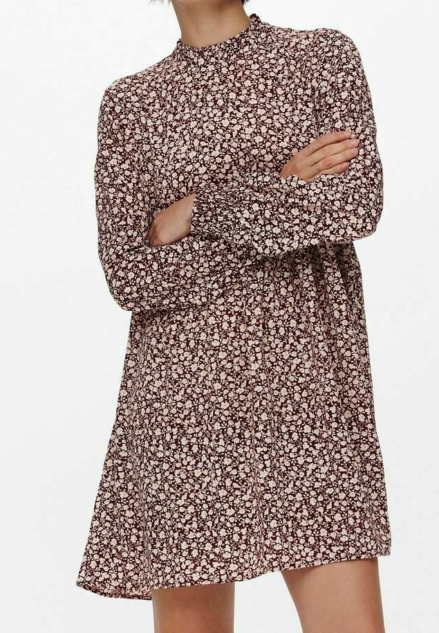 Femme portant une robe à manches longues et col montant avec un motif floral rose et marron, bras croisés, debout devant un fond blanc uni.