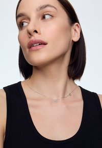 Srebrny naszyjnik z łańcuszka z delikatnym kwiatowym wisiorkiem, teksturowanymi akcentami i minimalistycznym wzornictwem, noszony na czarnej bezrękawnik.