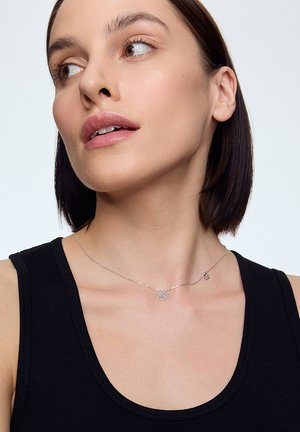 Srebrny naszyjnik z łańcuszka z delikatnym kwiatowym wisiorkiem, teksturowanymi akcentami i minimalistycznym wzornictwem, noszony na czarnej bezrękawnik.