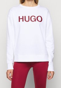 Vit sweatshirt med en framträdande vinröd "HUGO"-logotyp. Den har en avslappnad passform, långa ärmar och ribbad nederkant och muddar.